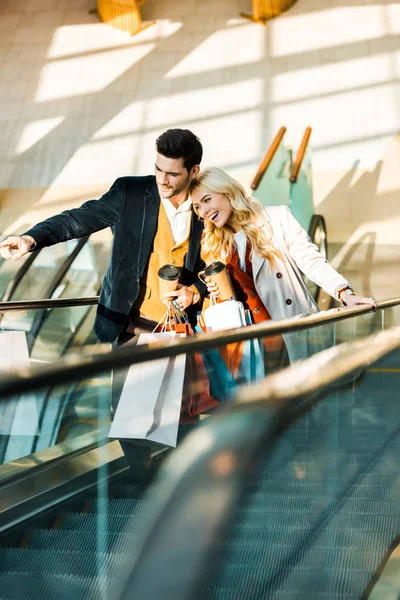 Felice uomo elegante con il caffè per andare a mostrare qualcosa alla ragazza con borse della spesa in piedi sulla scala mobile — Foto stock