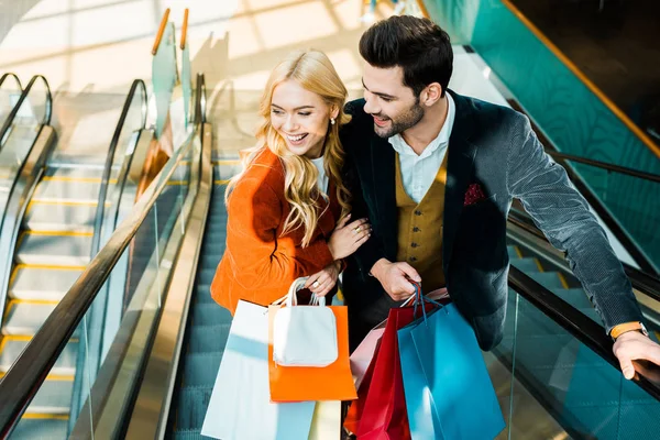 Elegant smiling couple with shopping bags standing on escalator — Stock Photo