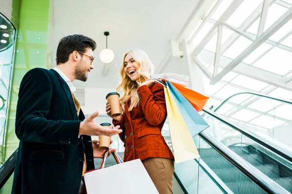 Beautiful stylish couple with shopping bags and coffee to go talking on escalator — Stock Photo