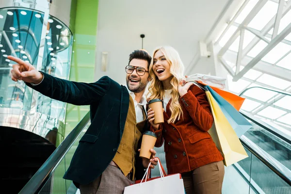 Fidanzato alla moda con il caffè per andare a mostrare qualcosa alla ragazza eccitata con le borse della spesa in piedi sulla scala mobile — Foto stock