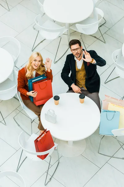 Overhead view of beautiful couple with shopping bags sitting in cafe and waving to camera — Stock Photo