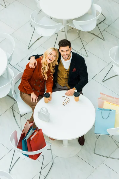 Над головой вид счастливой пары с мешками для покупок, обнимающих и сидящих в кафе — стоковое фото