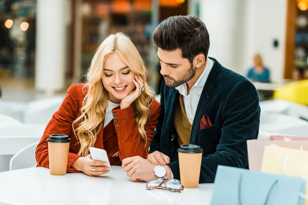 Sonriente pareja usando smartphone en cafetería con café para ir y bolsas de compras - foto de stock