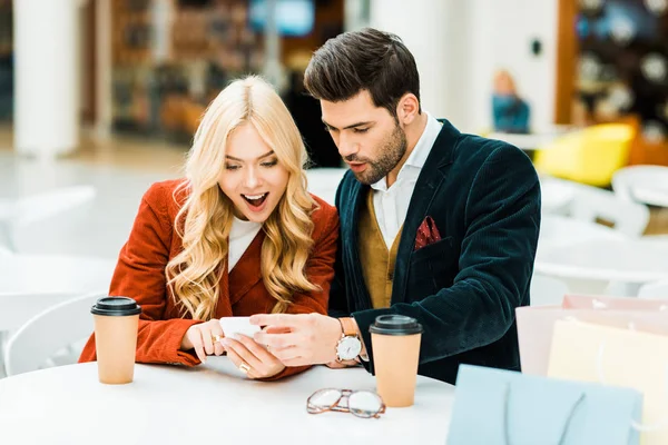 Casal animado usando smartphone no café com café para ir e sacos de compras — Fotografia de Stock