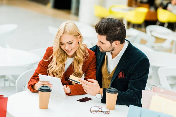 Улыбающаяся пара делает покупки онлайн с помощью кредитной карты и цифрового планшета, сидя в кафе в торговом центре — стоковое фото