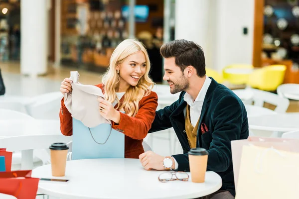 Happy stylish girl showing new clothes to smiling boyfriend in cafe in shopping mall — Stock Photo