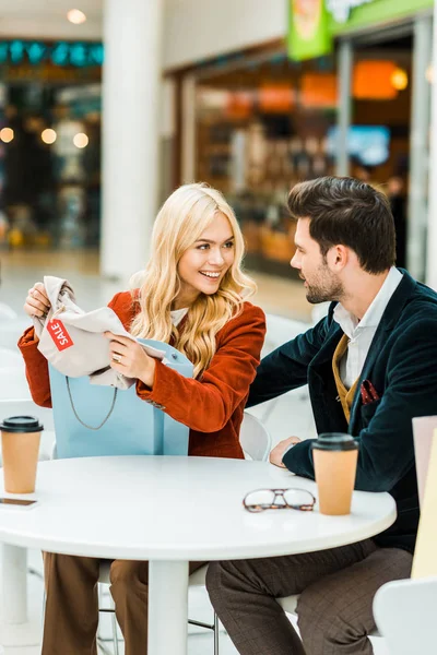 Beautiful blonde girl showing new clothes to boyfriend in cafe in shopping center — Stock Photo