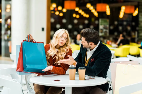 Mujer confundida mostrando bolsas de compras al hombre en la cafetería en el centro comercial - foto de stock
