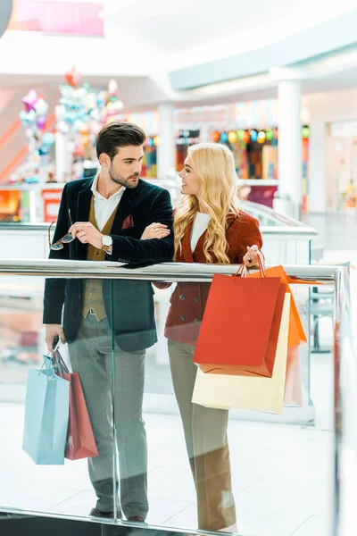 Jovem casal elegante segurando sacos de compras e olhando uns para os outros no shopping — Fotografia de Stock