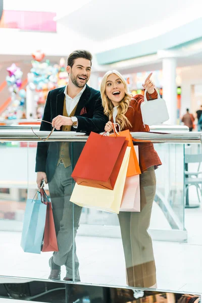 Pareja joven con bolsas de compras pasar tiempo en el centro comercial, mujer mostrando algo - foto de stock