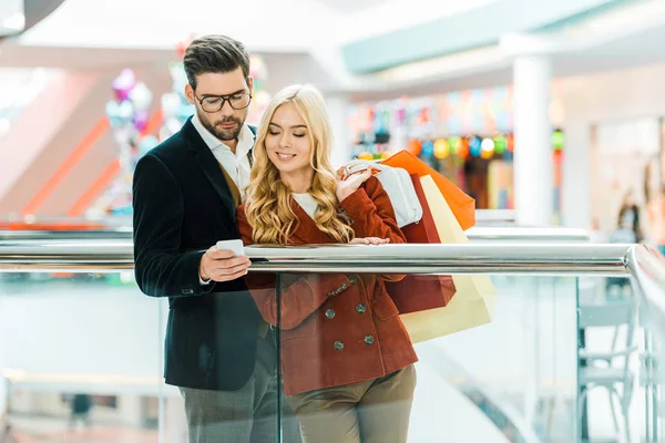 Pareja joven y elegante con bolsas de compras usando teléfono inteligente en el centro comercial - foto de stock