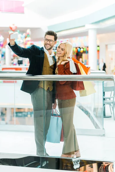 Smiling fashionable couple with shopping bags taking selfie in shopping mall — Stock Photo