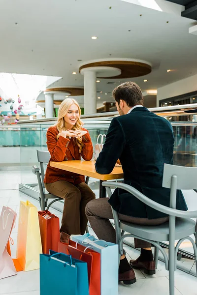 Hermosa pareja mirándose mientras se sienta con bolsas de compras en la cafetería en el centro comercial - foto de stock