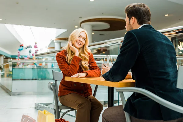 Счастливая пара смотрит друг на друга и сидит с сумками в кафе в торговом центре — стоковое фото