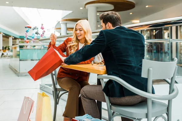 Hermosa mujer feliz mostrando algo en bolsa de compras a novio mientras está sentado en la cafetería en el centro comercial - foto de stock