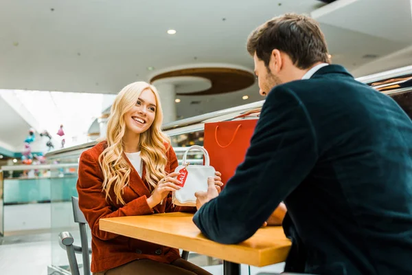 Smiling woman showing bag with sale tag to boyfriend in shopping mall — Stock Photo