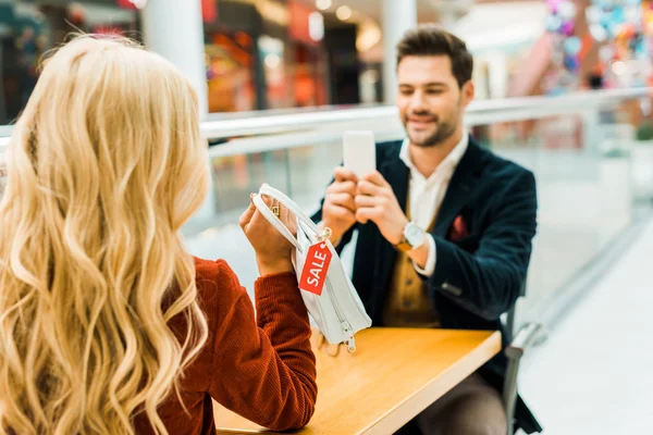 Stylish man taking photo of blonde girlfriend holding bag with sale tag — Stock Photo