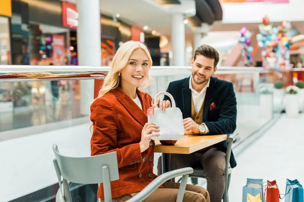 Hermosa shopaholic mostrando bolsa con etiqueta de venta en el centro comercial - foto de stock