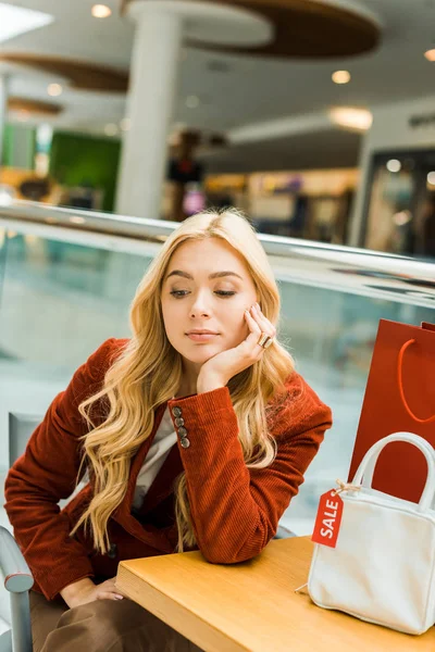 Upset blonde girl sitting in mall with one shopping bag and one bag with sale tag — Stock Photo