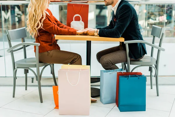 Vista recortada de la pareja cogida de la mano mientras se sienta con bolsas de compras en la cafetería en el centro comercial - foto de stock
