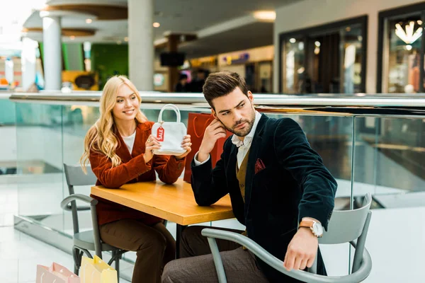 Female shopaholic showing bag with sale tag to upset boyfriend in shopping center — Stock Photo