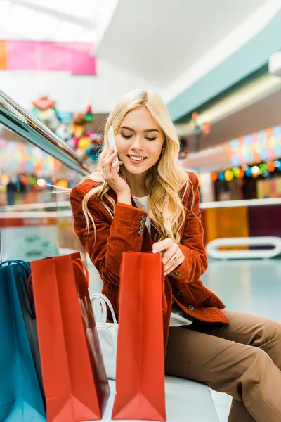 Atractiva mujer hablando en el teléfono inteligente y mirando en bolsas de compras - foto de stock