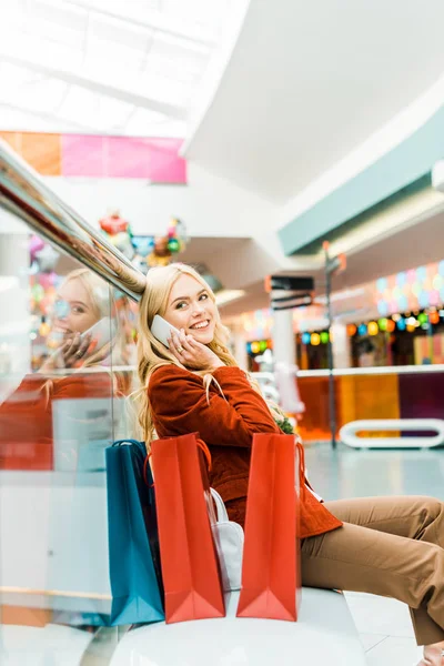 Schönes Mädchen, das auf dem Smartphone spricht und mit Einkaufstüten in Einkaufszentrum sitzt — Stockfoto