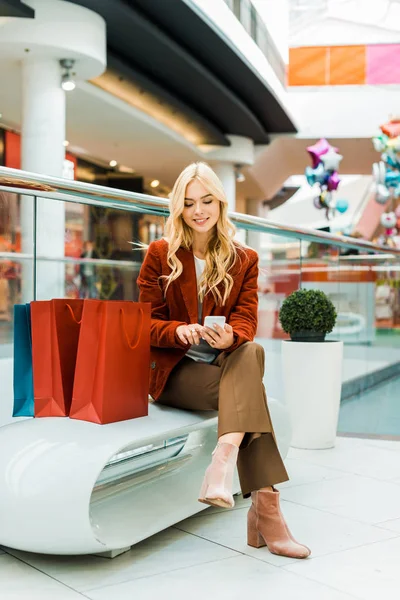 Hermosa mujer rubia usando teléfono inteligente y sentado con bolsas de compras en el centro comercial - foto de stock