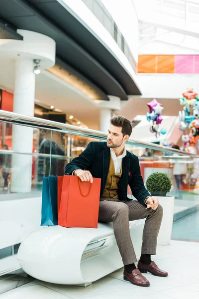 Fashionable man holding credit card and sitting with bags in shopping center — Stock Photo