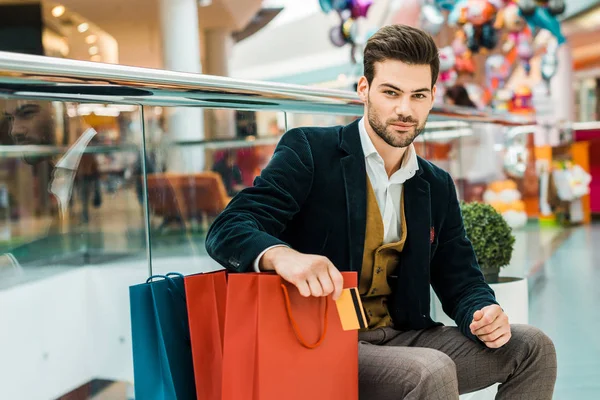 Elegante hombre con tarjeta de crédito y sentado con bolsas en el centro comercial - foto de stock