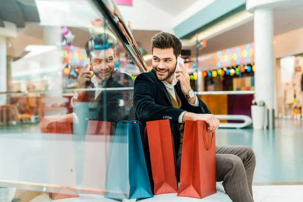 Homme souriant parlant sur smartphone tout en étant assis dans le centre commercial avec des sacs — Photo de stock