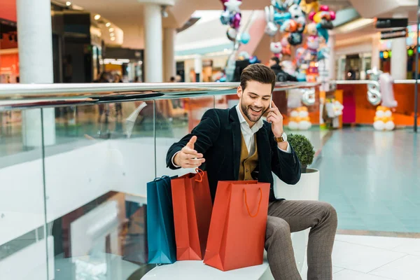 Bel homme à la mode avec des sacs à provisions parler sur smartphone tout en étant assis dans le centre commercial — Photo de stock