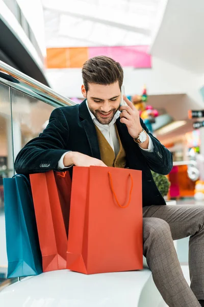 Стильный мужчина смотрит в сумки и разговаривает по смартфону, сидя в торговом центре — стоковое фото