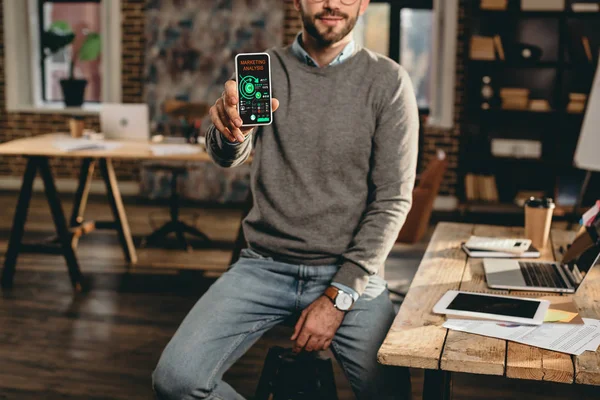 Vue recadrée d'un homme d'affaires occasionnel tenant un smartphone avec une application marketing analisys à l'écran dans un bureau loft — Photo de stock