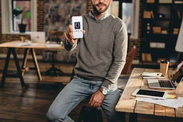 Vista recortada de hombre de negocios casual celebración de teléfono inteligente con aplicación uber en la pantalla en la oficina loft - foto de stock