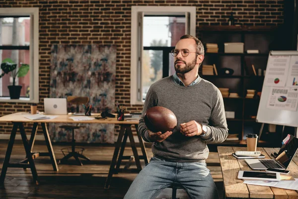 Hombre de negocios casual sosteniendo pelota de rugby y sentado en la oficina loft - foto de stock