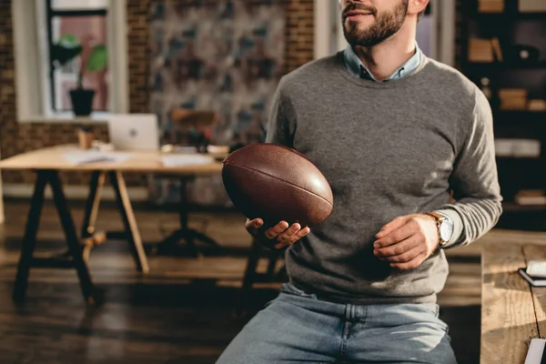 Cropped view of casual businessman holding rugby ball and sitting in loft office — Stock Photo