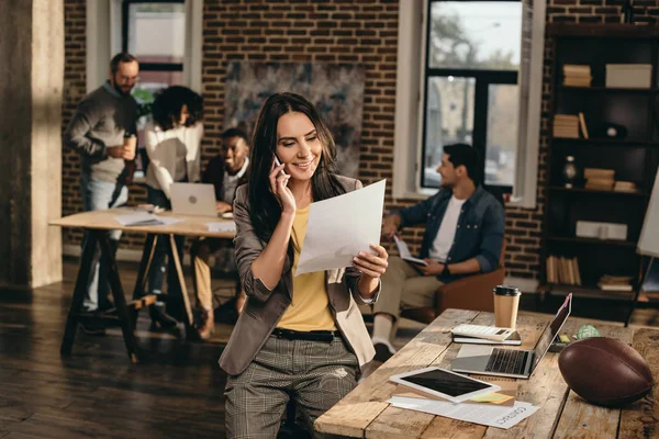 Femme d'affaires occasionnelle travaillant et parlant sur smartphone dans le bureau loft avec des collègues derrière — Photo de stock