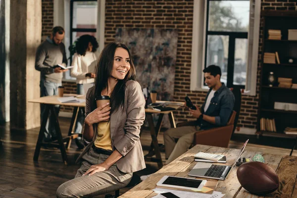 Усміхнена випадкова бізнес-леді тримає чашку кави в офісі лофт з колегами, що працюють позаду — стокове фото
