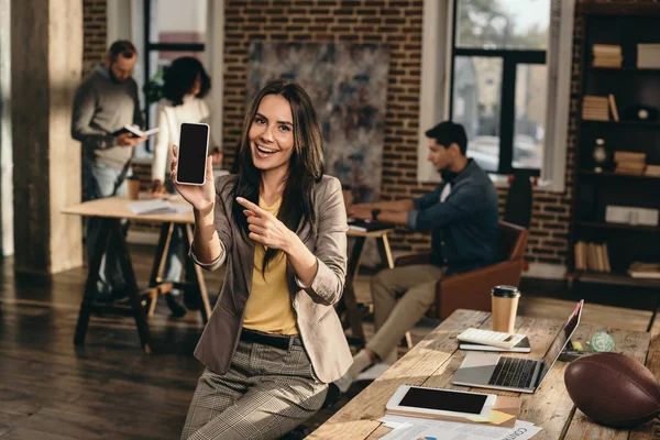 Sonriente mujer de negocios casual sosteniendo teléfono inteligente con pantalla en blanco en la oficina loft con colegas que trabajan detrás — Stock Photo