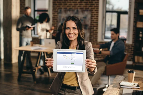 Gelegenheitsunternehmerin hält Tablet mit Facebook-Website auf dem Bildschirm im Loft-Büro mit Kollegen, die dahinter arbeiten — Stockfoto