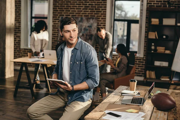 Young man holding tablet in modern loft office with colleagues working on background — Stock Photo