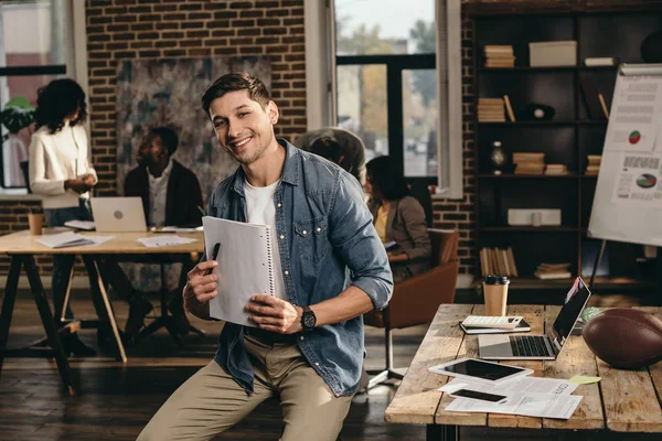 Smiling young man looking at camera, holding pen and journal in modern loft office with colleagues working on background — Stock Photo