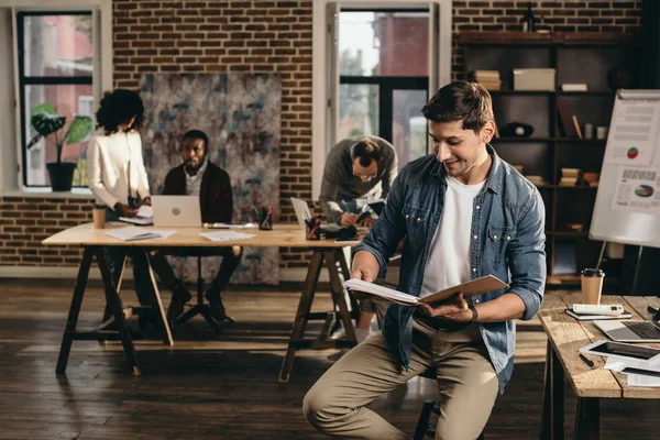 Young man looking at journal in modern loft office with colleagues working on background — Stock Photo