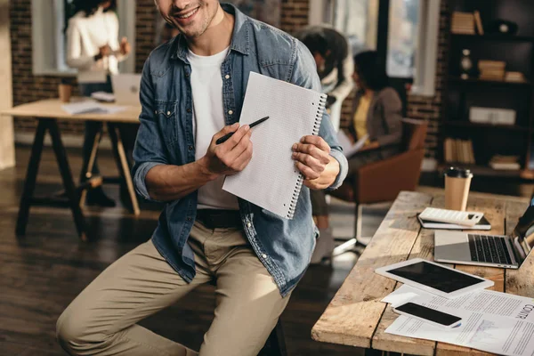 Cropped view of young man pointing with pen on journal with colleagues on background in modern loft office — Stock Photo