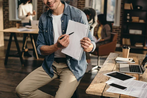 Vista recortada de hombre joven señalando con pluma en el diario con colegas en el fondo en la oficina loft moderno - foto de stock