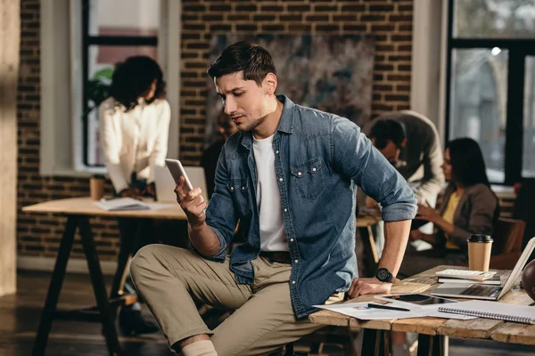 Young man using smartphone in modern loft office with working colleagues on background — Stock Photo