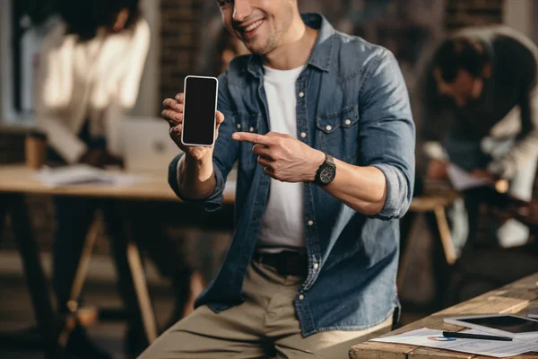 Vista ritagliata di giovane uomo sorridente che punta il dito sullo smartphone in un moderno ufficio loft con colleghi sullo sfondo — Foto stock