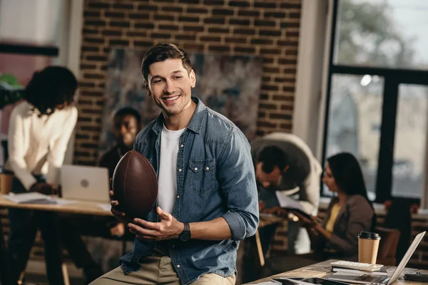 Smiling young man holding rugby ball in modern loft office with colleagues on background — Stock Photo