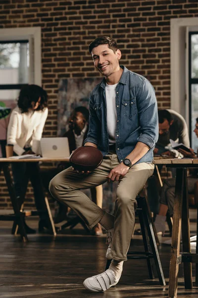 Lächelnder junger Mann mit Rugby-Ball im modernen Loft-Büro mit Kollegen im Hintergrund — Stockfoto